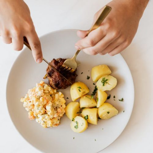 Top view of crop unrecognizable male with cutleries slicing delicious beef cutlet garnished with boiled potatoes and parsley and portion of Russian meat salad on plate placed on white table